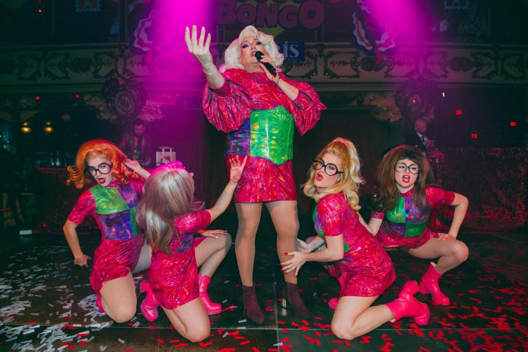 Drag queen y azafatas de Loco Bongo posando en el escenario bajo luces rosas